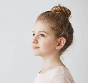 Close up of good-looking little girl with blonde hair in bun hairstyle, standing in three quarters, looking aside with smile on her face